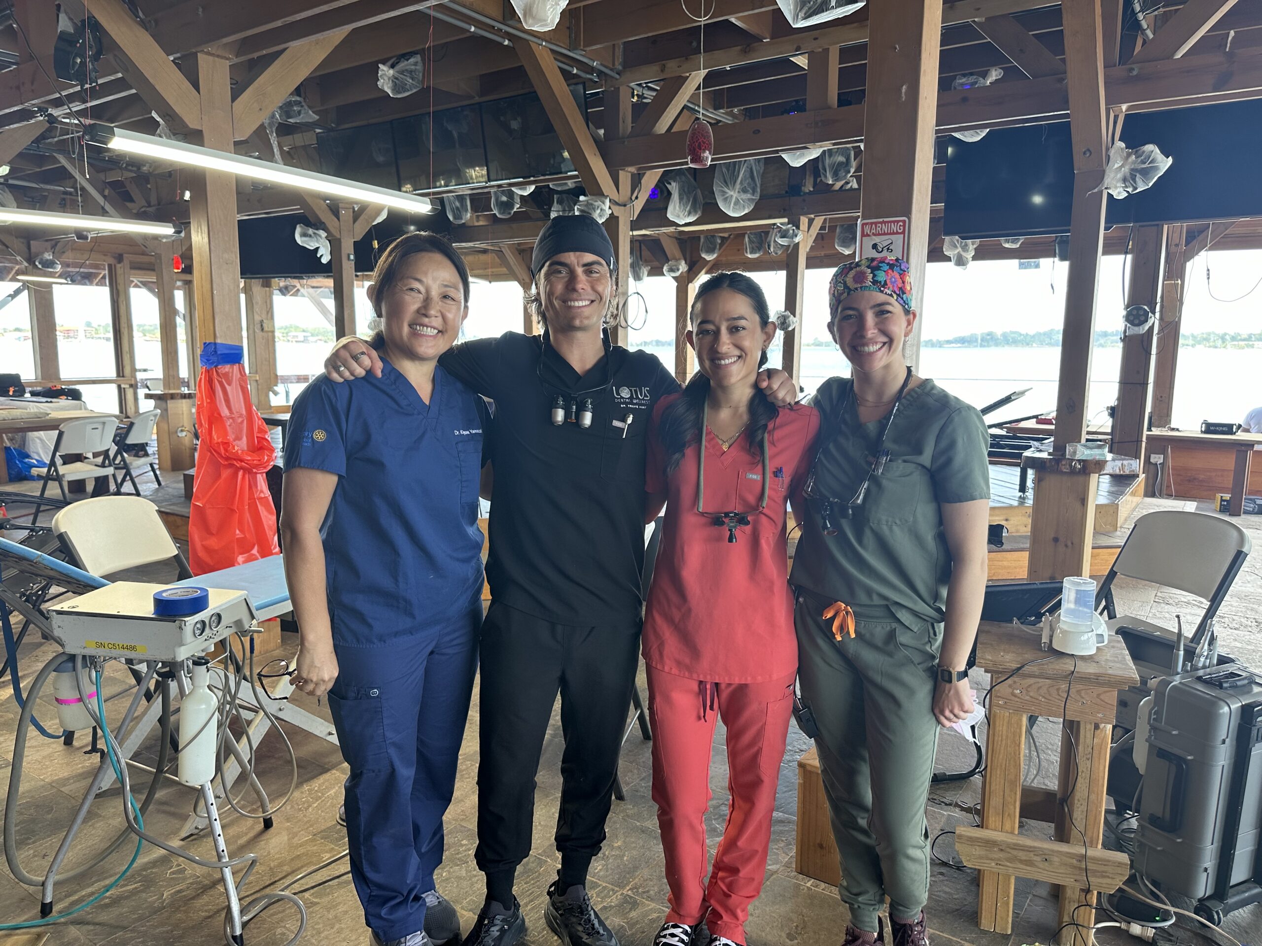 Dr. Kiyomi Yamazaki, Dr. Travis Hunt, Ashley Affholter and Dr. Jennifer Stone in scrubs, smiling among portable dental chairs and equipment in the open air clinic at Tropical Arts, with the bay behind them