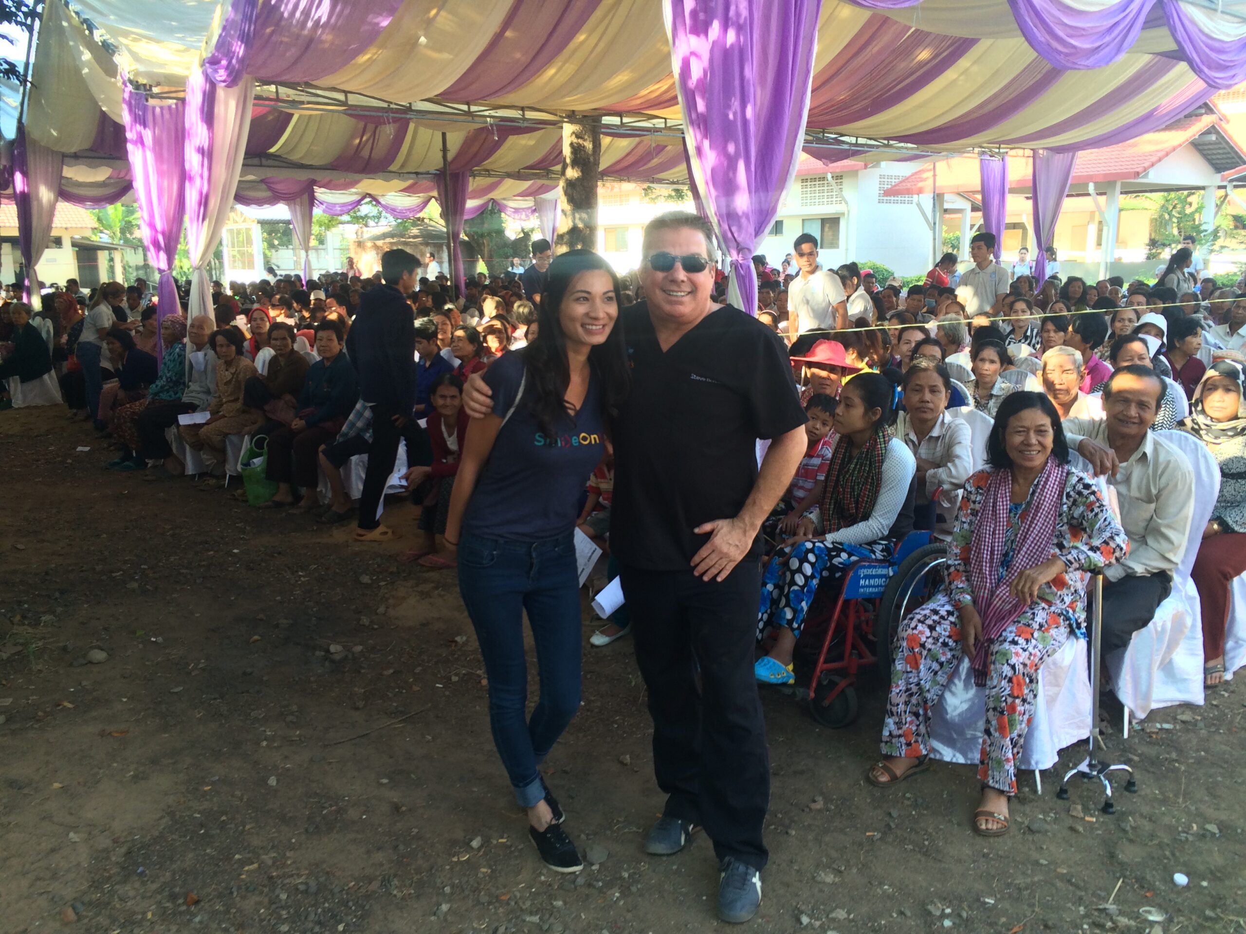 Steve Bender and a SmileOnU volunteer stand in front of hundreds of patients seated under a purple and white tent, waiting for the free clinic in Siem Reap, Cambodia