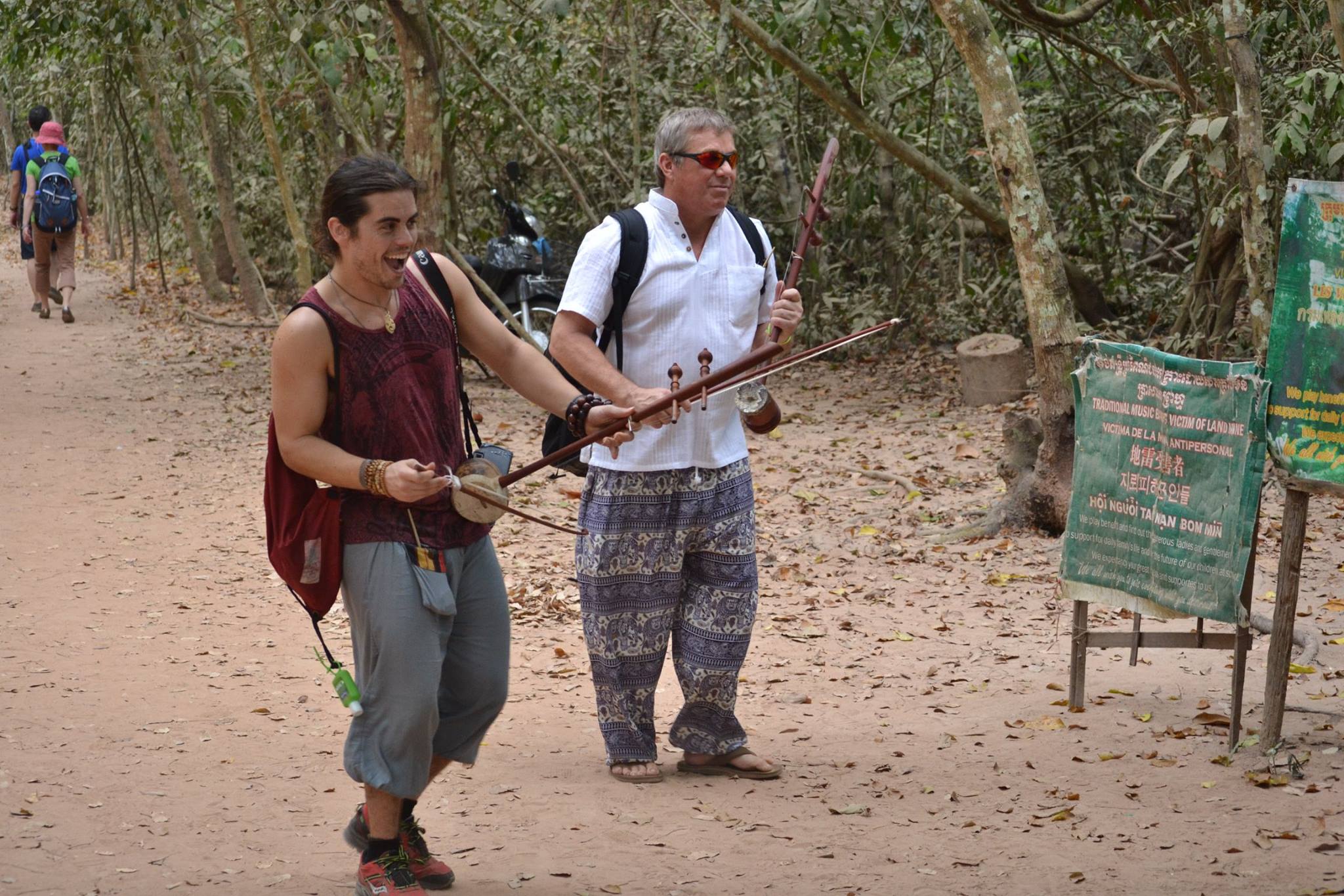 Steve Bender and a young volunteer holding traditional Khmer two string fiddles beside a sign for a music band of landmine victims near Angkor