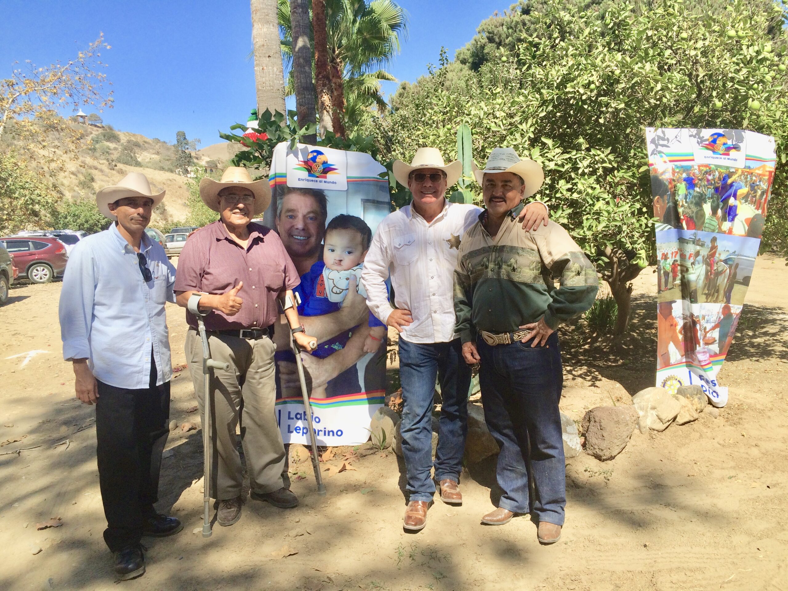 Steve Bender in a cowboy hat with Rosarito Rotarians beside a banner of Steve holding baby Christopher before his cleft lip surgery