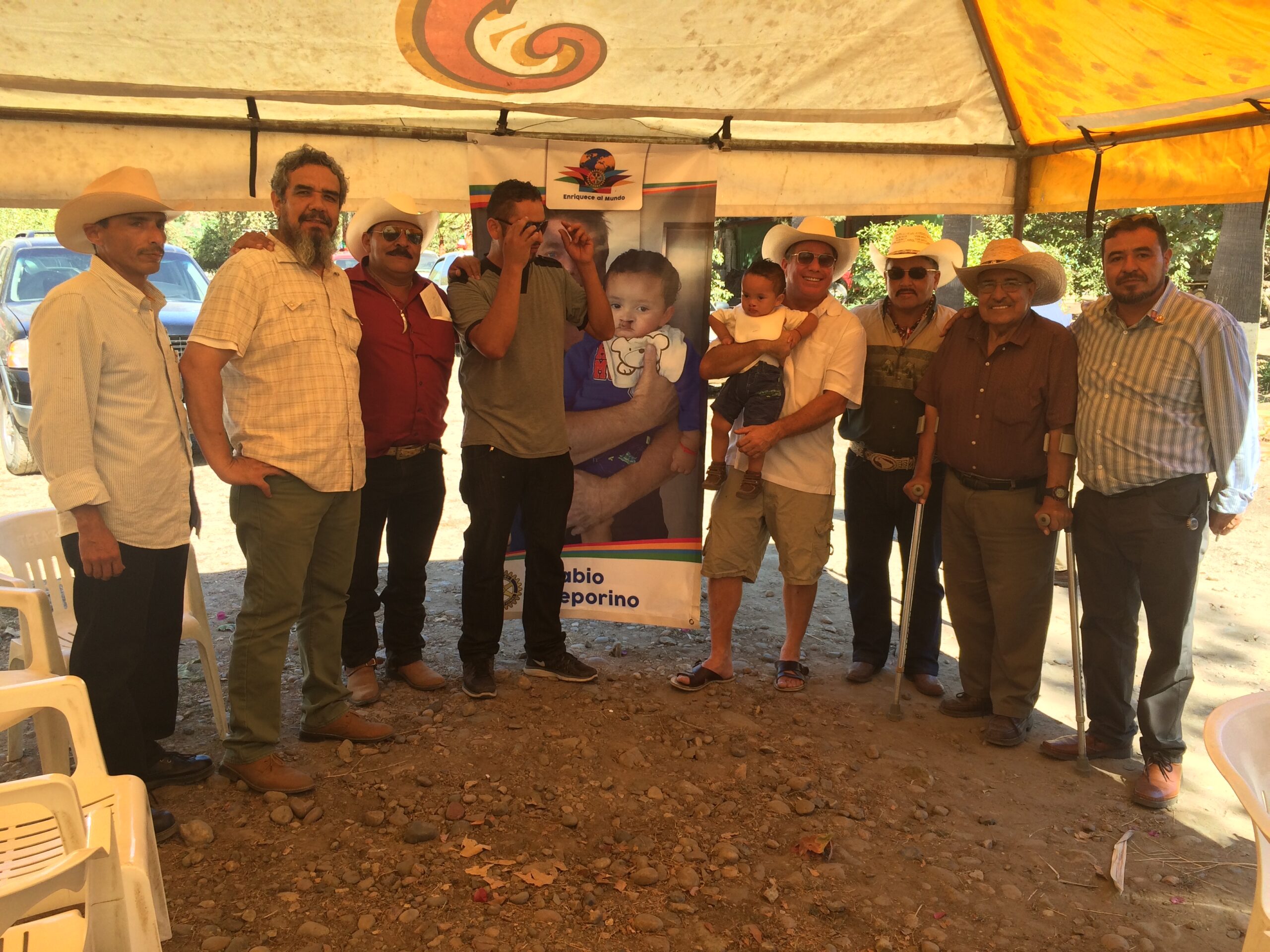 Steve Bender holding a little boy under a tent with Rosarito Rotarians beside the Labio Leporino banner