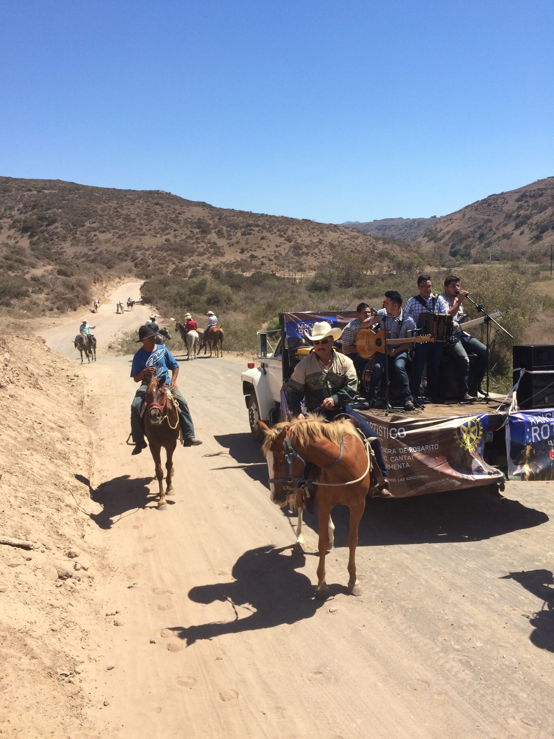 Riders on horseback beside a truck carrying a norteño band with a Rotary banner on a dirt road in the hills