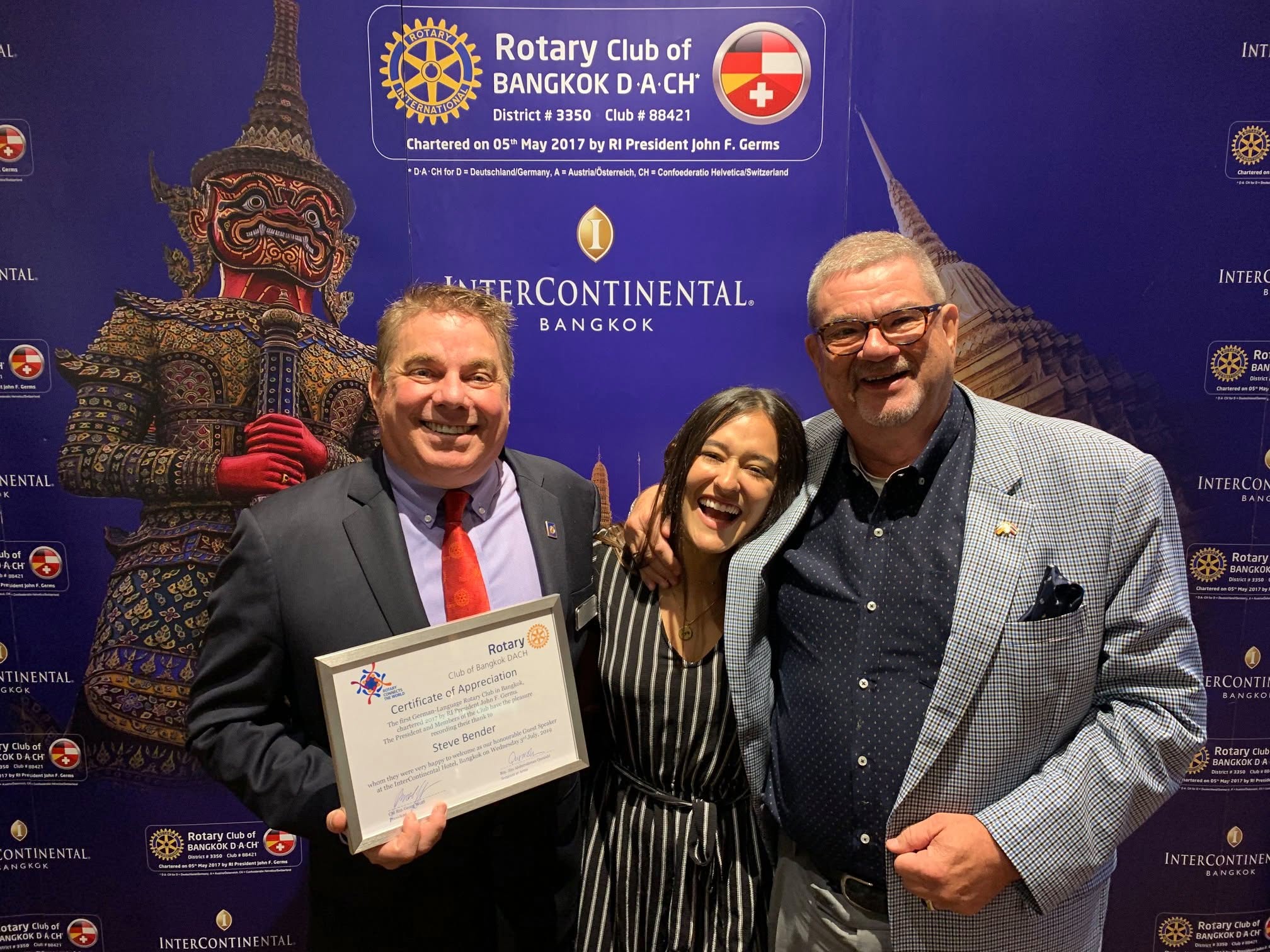 Steve Bender holding a framed Certificate of Appreciation from the Rotary Club of Bangkok DACH, with Ashley Affholter and a club member at the InterContinental Bangkok