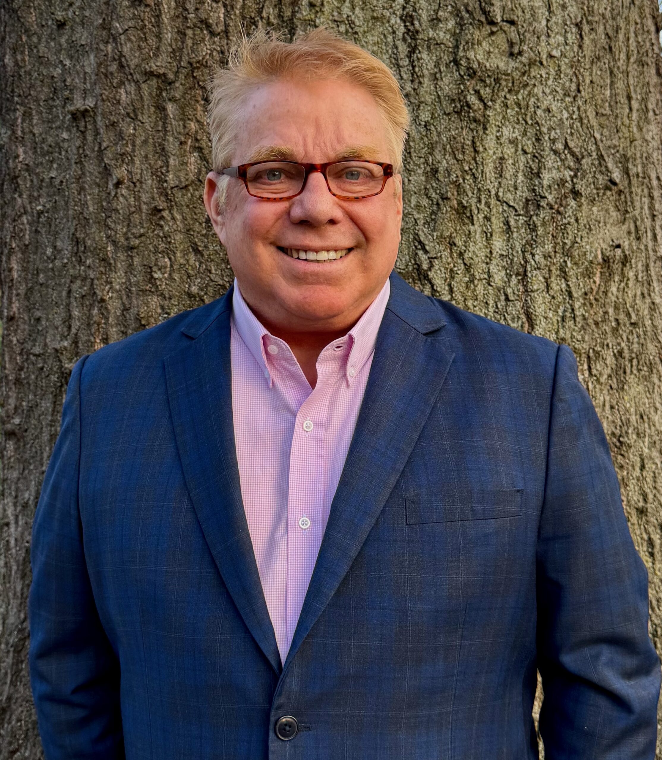 Steve Bender in a navy blazer and pink shirt, smiling in front of a large tree trunk