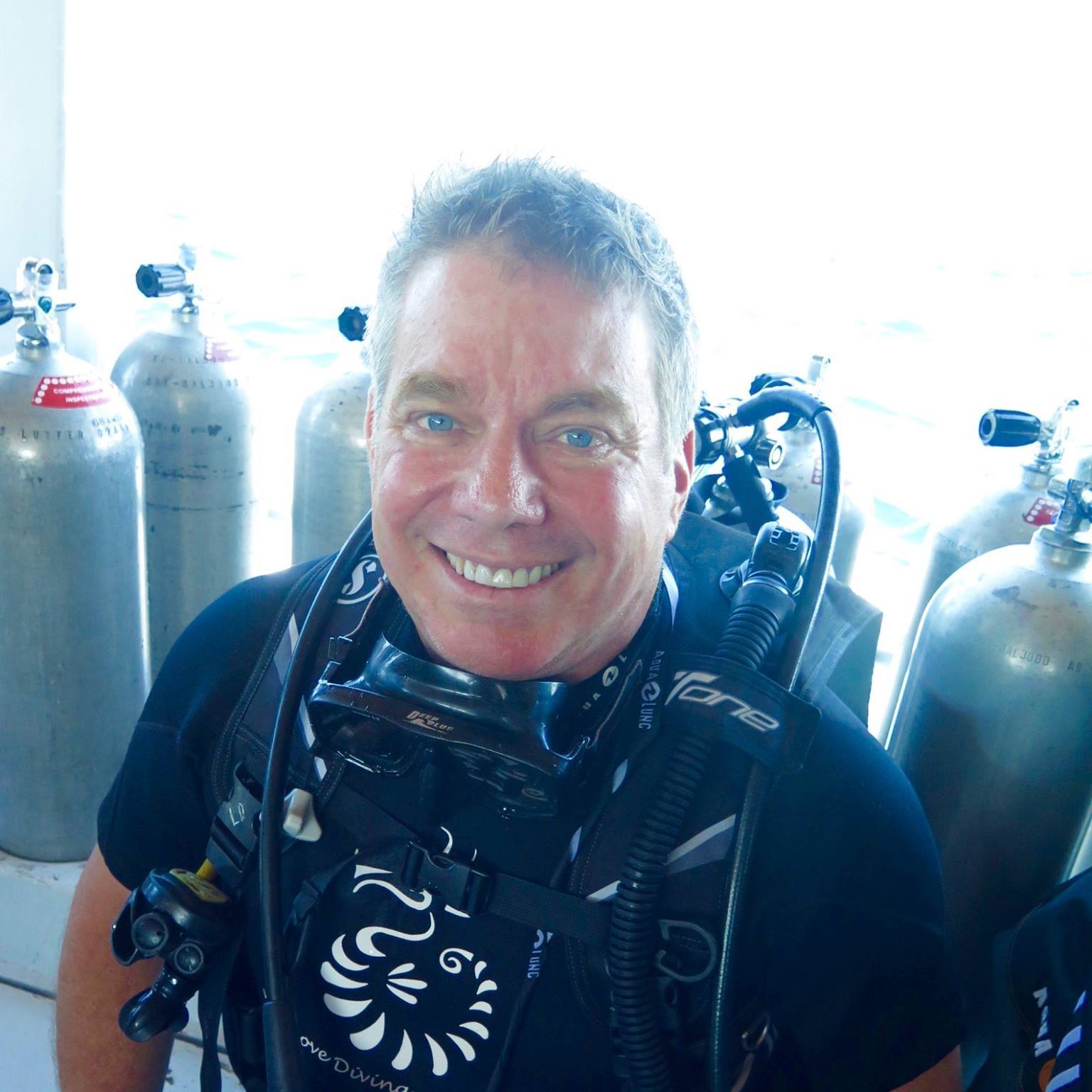 Steve Bender smiling in scuba gear with a dive mask around his neck, beside rows of tanks on a dive boat